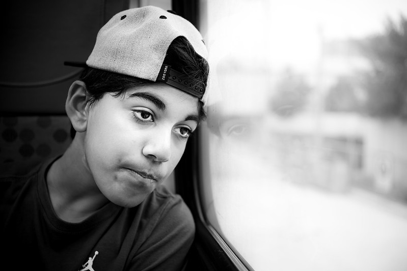 A morose boy in a hat looks out a vehicle window.