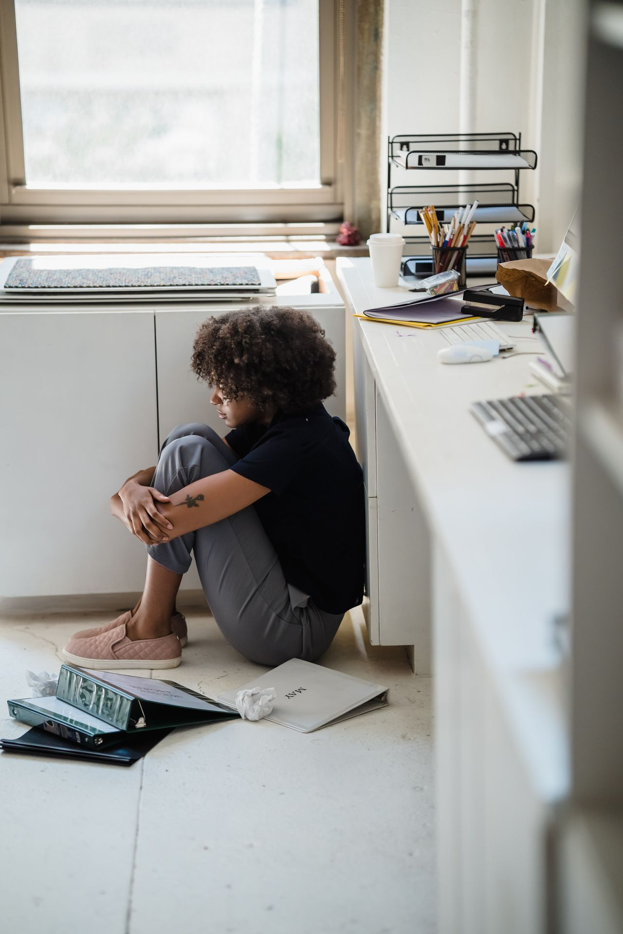 A young woman crouched in the corner of an office, surrounded by documents and binders.