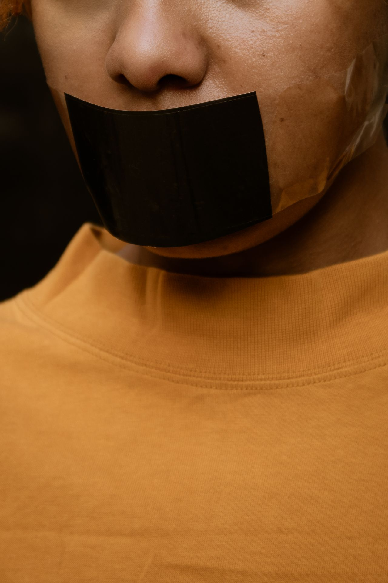 Woman of color wearing a yellow shirt and with black tape across their mouth