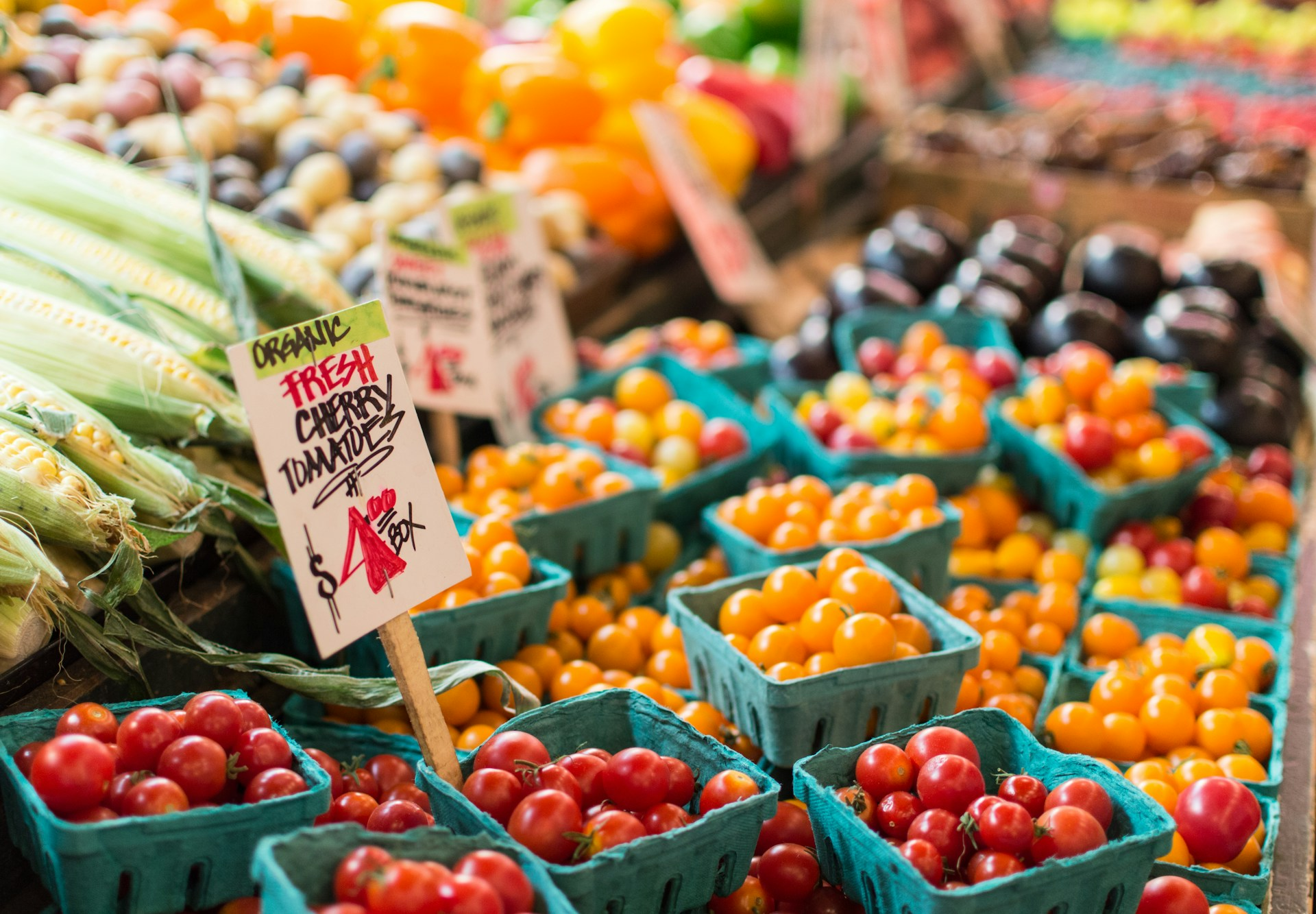 Vegetable market with a focus on boxes of cherry tomatoes and partially husked corn on the cob