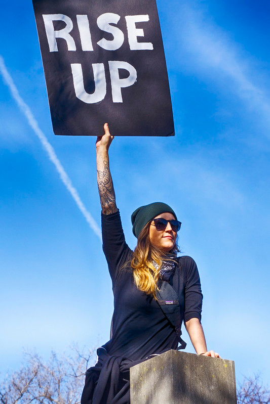 Woman in a black shirt holding up a protest sign reading RISE UP